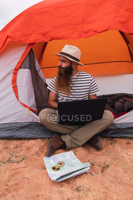 Muchacho barbudo atractivo usando el ordenador portátil y mirando hacia otro lado mientras está sentado cerca de la tienda y el mapa con brújula - foto de stock