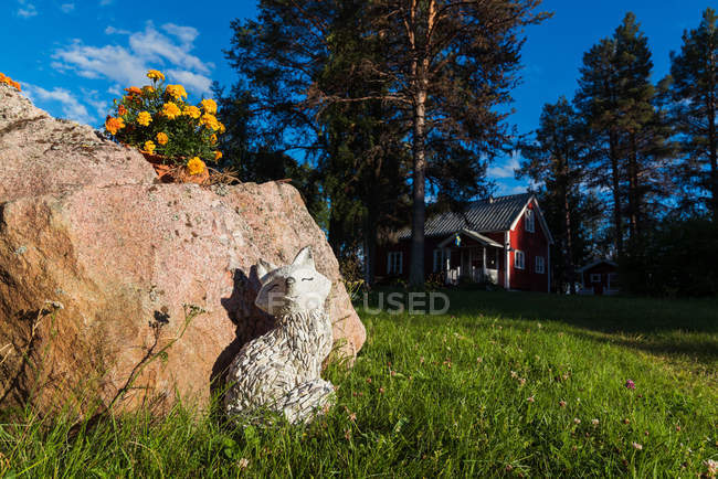 Statue of dog made of white stone near big rock with flowers with wooden house among trees on background — Stock Photo