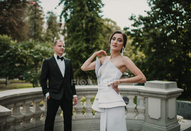 Charming young woman in beautiful wedding dress posing for camera while standing near groom on terrace of luxury building — Stock Photo