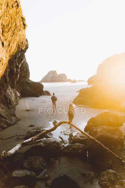 Back view of young man and woman walking on wet sand near stone cliffs and sea during sunset on beach in California — Stock Photo