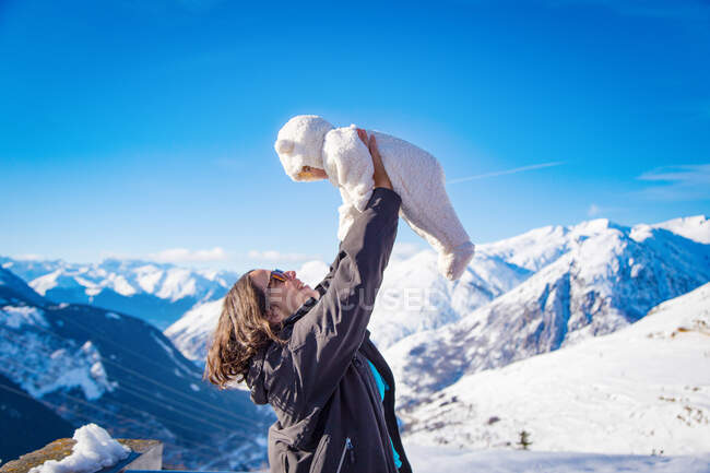 Side view of excited young female smiling and lifting cute baby while standing on background of amazing snowy mountains and clear blue sky — Stock Photo