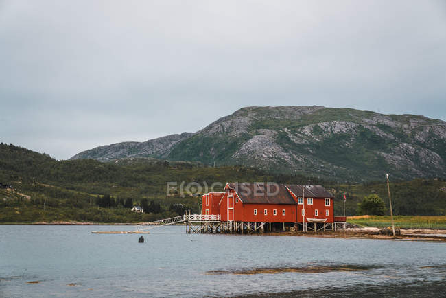 Cais com casa de madeira vermelha em pé no lago ondulante limpo no fundo das montanhas — Fotografia de Stock