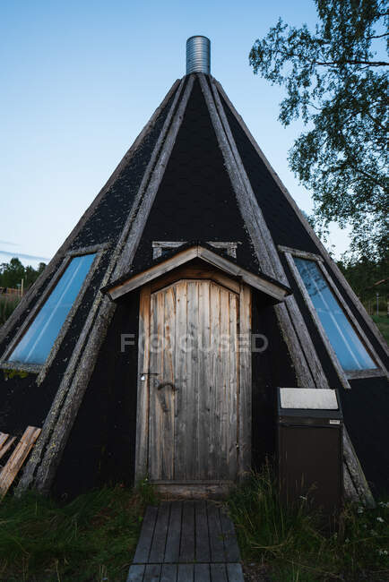 Casa en forma de triángulo con puerta de madera de pie en el bosque cerca del árbol en el fondo del cielo azul sin nubes - foto de stock