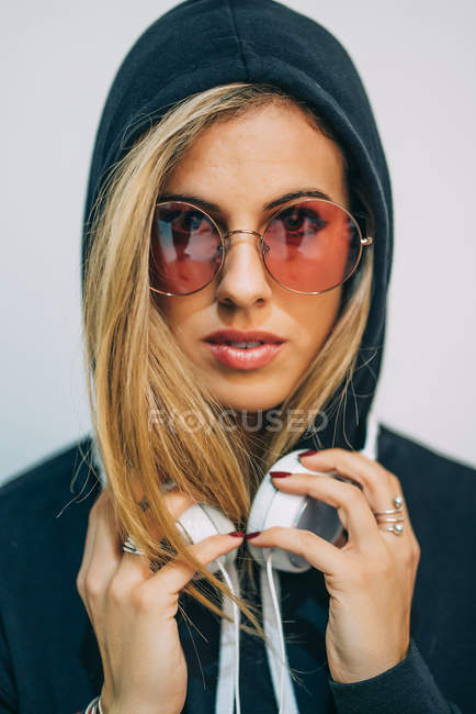 Mulher loira jovem com capuz preto e óculos de sol redondos com fones de ouvido no pescoço olhando para a câmera no fundo branco — Fotografia de Stock