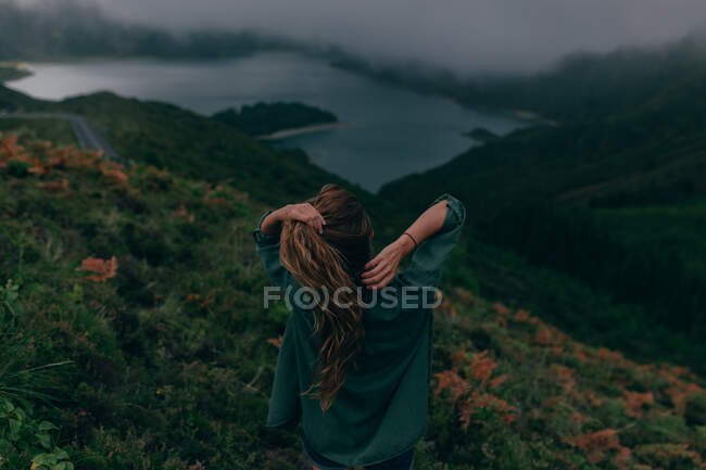 Вид сзади на молодую женщину, стоящую на холме с зеленой травой и смотрящую на красивое озеро внизу — стоковое фото