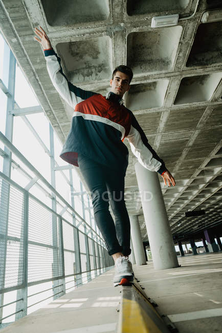 Jovem bonito homem no esporte jaqueta andando sobre ferro ferroviário em edifício espaçoso com pilares — Fotografia de Stock
