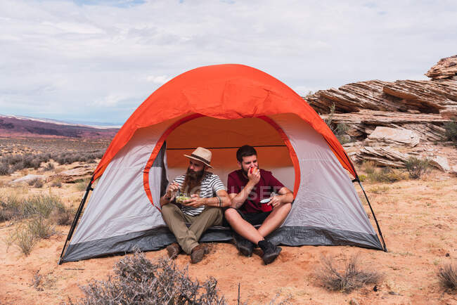Два друга-мужчины улыбаются и сидят рядом с палаткой вместе и едят — стоковое фото
