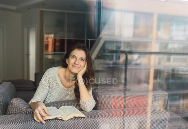 Прекрасна доросла жінка в елегантному одязі посміхається і озирається геть, лежачи на зручному дивані з цікавою книгою — стокове фото