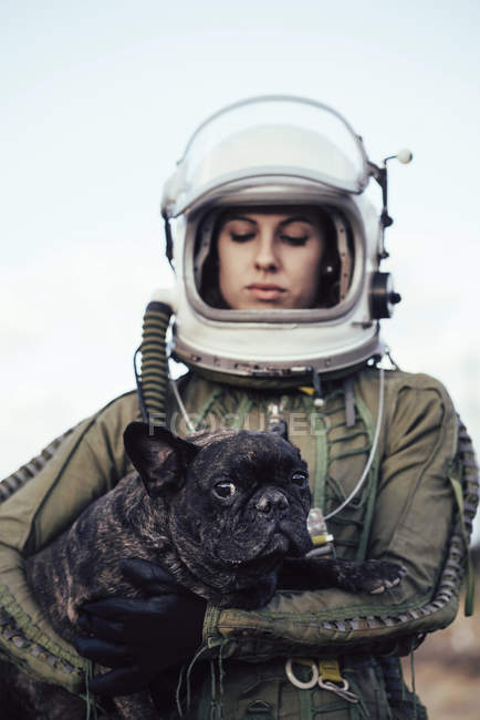 Улыбающаяся девушка в старом космическом шлеме и скафандре, держащая собаку на природе — стоковое фото
