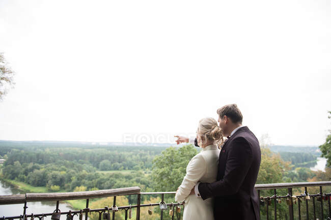 Back view of embracing elegant bride and groom standing on terrace with padlocks on fence and exploring nature views — Stock Photo