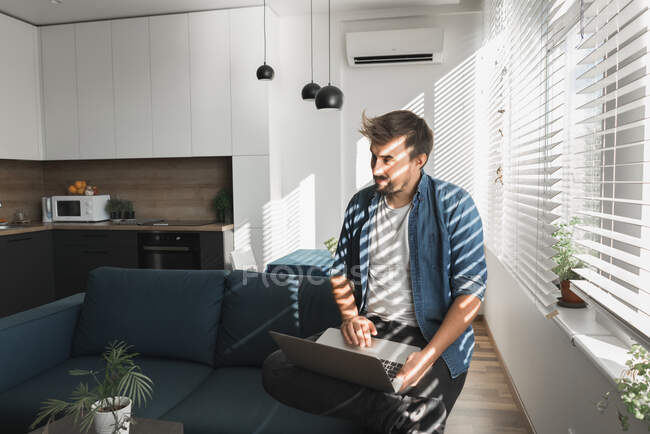 Bärtiger Mann surft auf modernem Laptop, während er auf bequemer Couch im stilvollen Wohnzimmer sitzt — Stockfoto