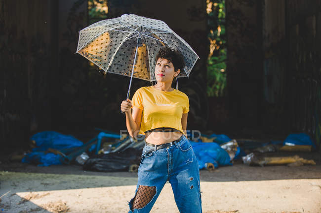 Nachdenkliche junge Frau in stylischem Outfit posiert mit durchsichtigem Regenschirm, während sie an sonnigen Tagen auf der Straße steht — Stockfoto