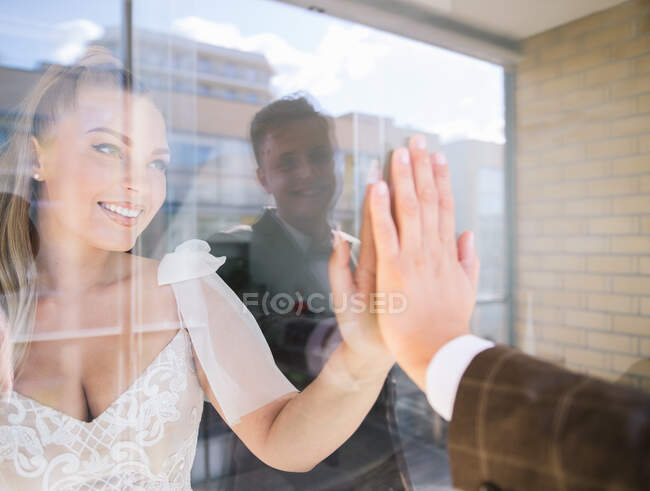 Cheerful newlywed couple touching hands while standing at glass — Stock Photo