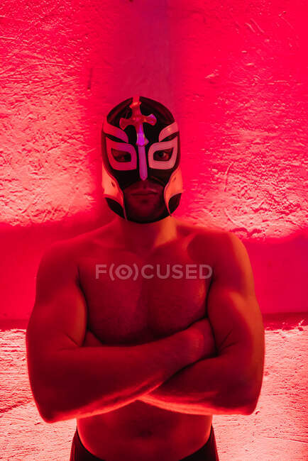 Shirtless muscular homem vestindo máscara de lutador e de pé com os braços cruzados em luz vermelha — Fotografia de Stock
