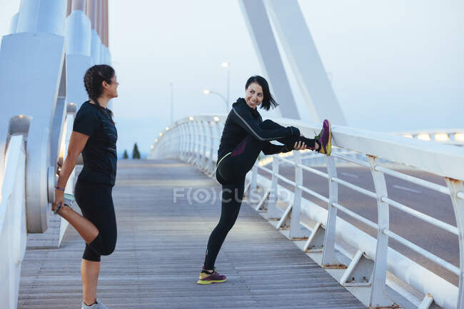 Вид сбоку улыбающихся женщин в спортивной одежде, разогревающихся перед тренировками на современном городском мосту — стоковое фото