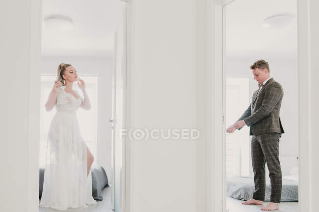 Hermosa mujer joven en vestido blanco y chico guapo en traje elegante preparándose para la ceremonia de boda en diferentes habitaciones de hotel - foto de stock