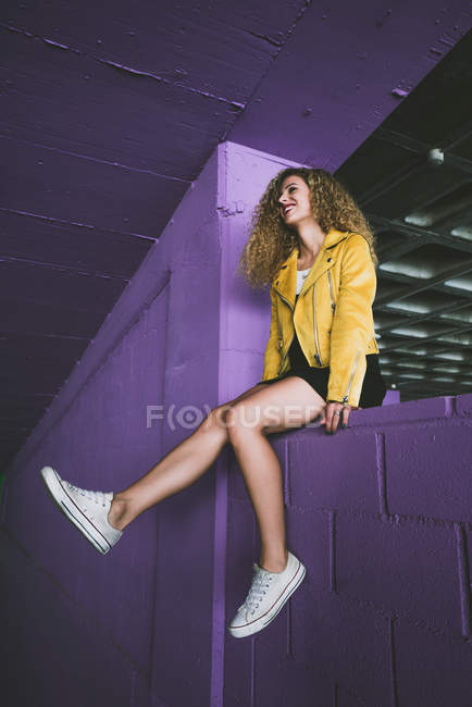 Élégante blonde bouclée en baskets et veste jaune assise sur le mur violet et riant — Photo de stock