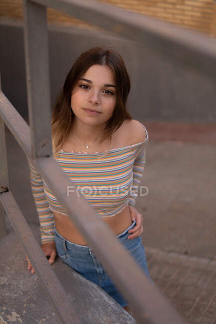 D'en haut de adorable jeune femme en tenue décontractée debout sur la rue derrière la clôture en regardant la caméra — Photo de stock