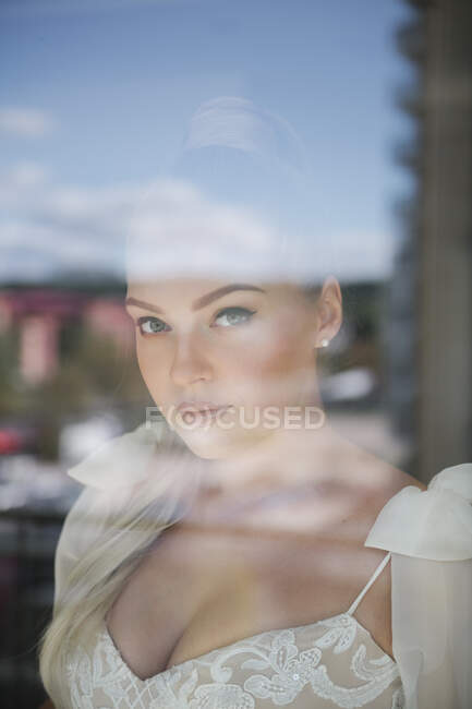 Mulher atraente em vestido de noiva branco em pé na janela e olhando para a câmera — Fotografia de Stock