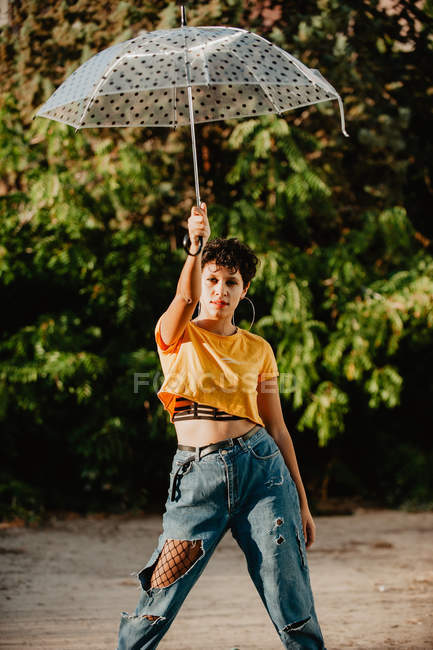 Привлекательная молодая женщина в стильной повседневной одежде смотрит в камеру, держа зонтик и стоя на улице возле кустов — стоковое фото