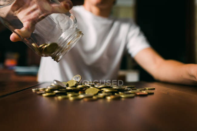 Joven anónimo derramando pequeñas monedas de tarro de vidrio en la mesa de madera - foto de stock