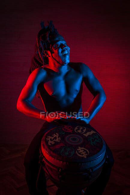 Jovem homem rastafarian africano gosta de ensaiar e joga tam tam tam, iluminação colorida vermelho e azul — Fotografia de Stock