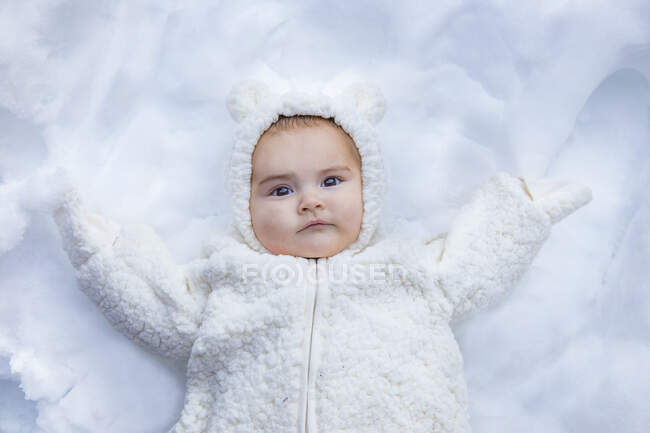 Сверху снимок сладкого малыша в теплой одежде, лежащего на белом снегу и смотрящего в камеру — стоковое фото