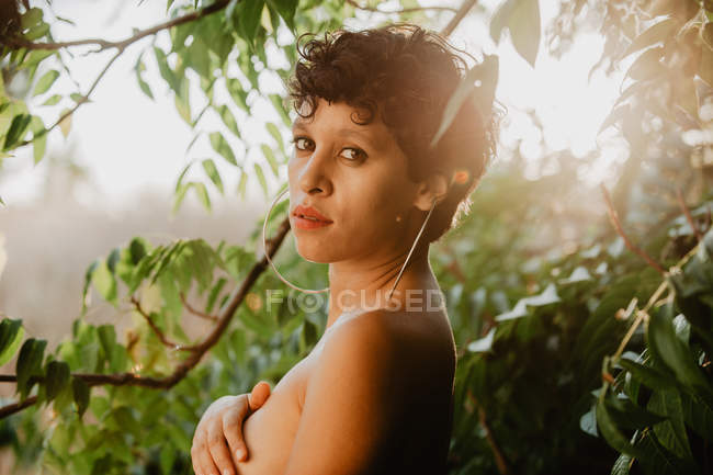 Portrait de brune sensuelle aux cheveux courts debout dans la brume dans la végétation verte avec la lumière du soleil — Photo de stock