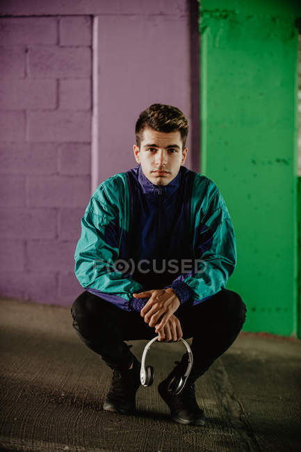 Young man in sportswear with headphones crouching against colorful wall — Stock Photo