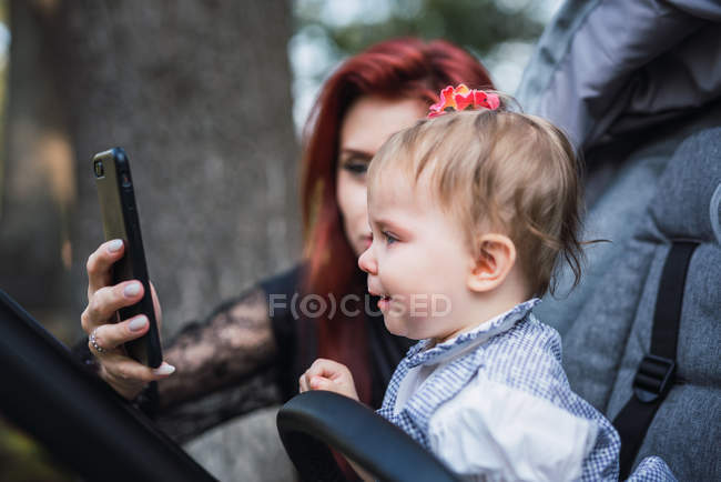 Mother showing smartphone to displeased baby in stroller — Stock Photo