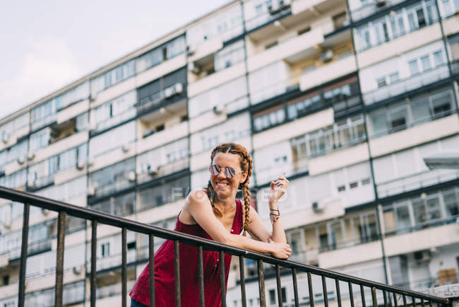 Руда молода жінка з косами і сонцезахисними окулярами курить проти житлового будинку — стокове фото