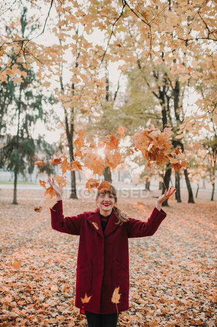 Stilvolle Frau in rotem Mantel wirft buntes Laub in Park und lacht — Stockfoto