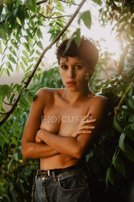 Чувственная молодая женщина, стоящая топлесс и покрывающая грудь руками в зеленом лесу — стоковое фото