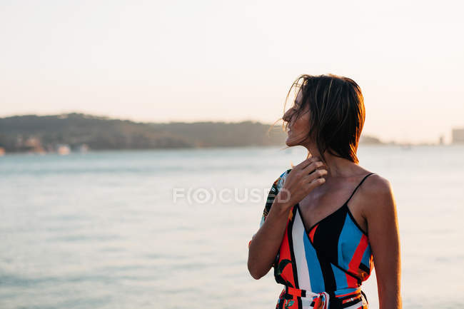 Thoughtful elegant woman standing on shore at sunset — Stock Photo