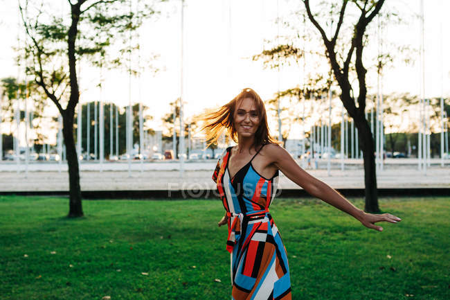 Joyeux brune élégante en robe tournant autour sur la prairie verte dans le parc urbain — Photo de stock