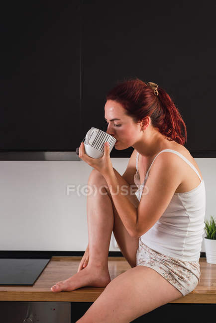 Woman sitting on kitchen counter and drinking coffee in kitchen — Stock Photo