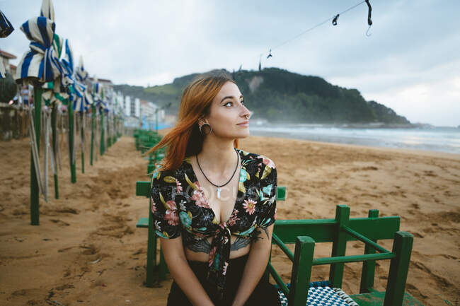 Attraente giovane donna sorridente e distogliendo lo sguardo mentre si siede sulla spiaggia di sabbia vicino al mare incredibile a Zarautz, Spagna — Foto stock