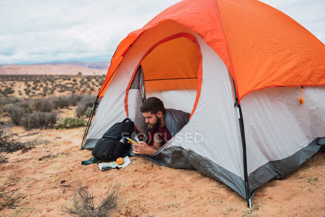 Cortar hombre barbudo comer manzana fresca y navegar por el teléfono inteligente moderno mientras está acostado en la tienda durante el camping en el desierto - foto de stock