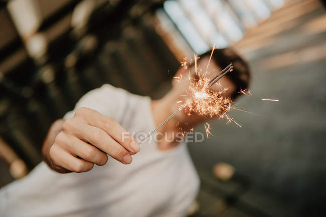 Молодой красивый мужчина в белой футболке стоит внутри здания и держит горящий бенгальский свет в руке — стоковое фото