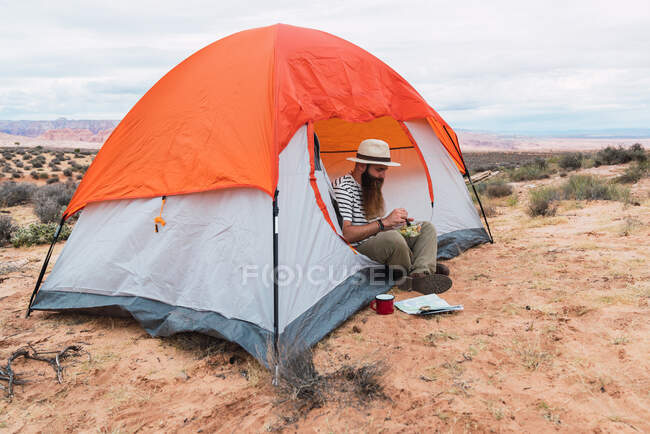 Homme manger de la salade et boire une boisson chaude tout en étant assis sur un sol sablonneux près de la carte et boussole pendant le camping dans le désert — Photo de stock