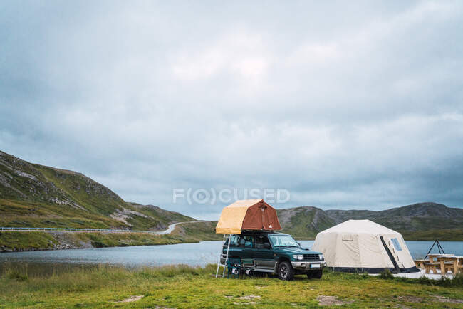 Автомобиль и палатка размещены на зеленом берегу синего тихого озера на фоне гор и облачного неба — стоковое фото