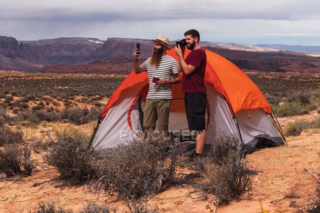 Two handsome men standing near tent and making pictures and videos of amazing desert while travelling together — Stock Photo