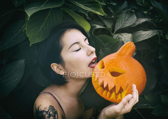 Side view of young brunette woman with tattoo and short hair standing near lush bush with closed eyes and holding decorative Jack O'Lantern in hands — Stock Photo