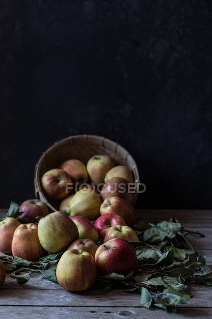 Свежие спелые яблоки и листья с корзиной на деревенском деревянном столе — стоковое фото