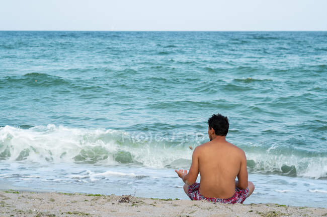 Vista trasera del macho en pantalones cortos sentados en la orilla arenosa cerca del magnífico mar ondulante en Tylenovo, Bulgaria - foto de stock