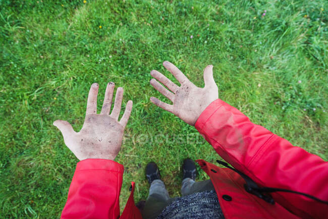 Ausschnitt von oben Ansicht einer Person in roter Jacke, die auf grünem Gras steht und mit Schmutz bedeckte Handflächen zeigt — Stockfoto