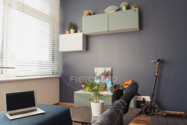 Piernas de los cultivos acostado en una pequeña mesa cerca de la computadora portátil con pantalla en blanco en la habitación elegante en apartamento moderno - foto de stock