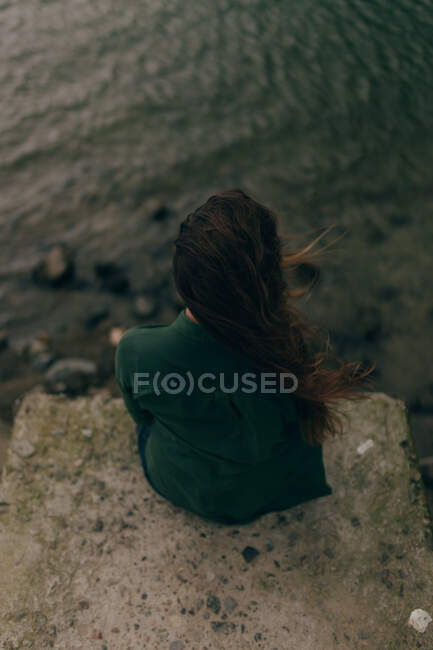 Вид сзади на молодую женщину с темными волосами в зеленой рубашке, сидящую на каменной скале с мелким озером внизу — стоковое фото