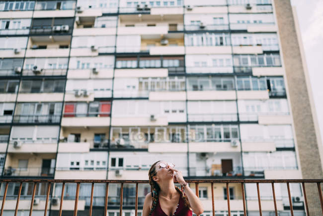 Стильная рыжая девушка с косичками и солнцезащитными очками, курящая против жилого дома — стоковое фото