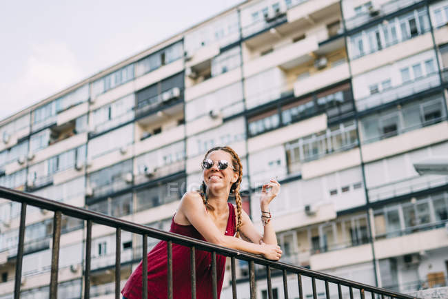 Red-haired young woman with braids and sunglasses smoking against residential building — Stock Photo